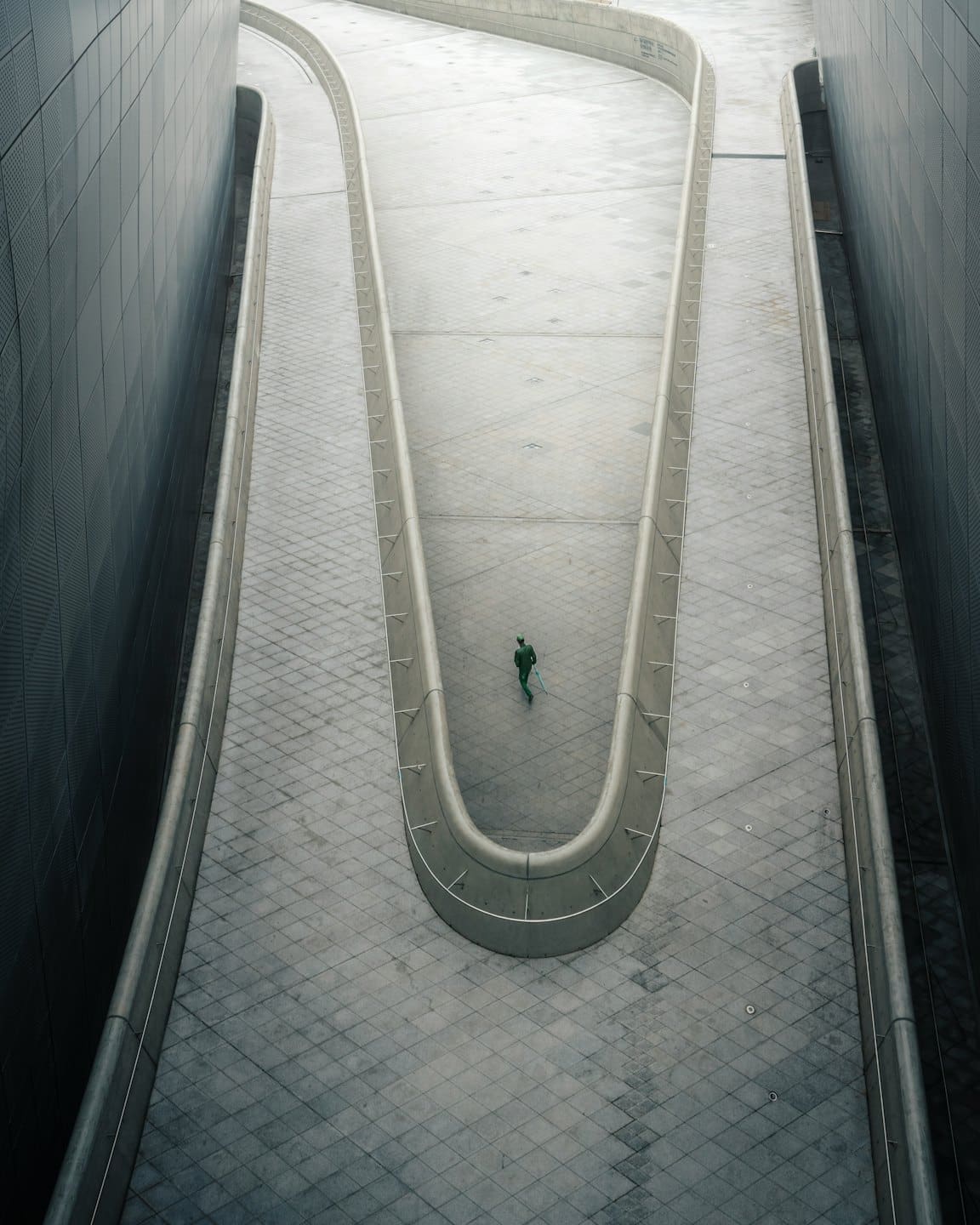Person in green walks a concrete path between textured walls.